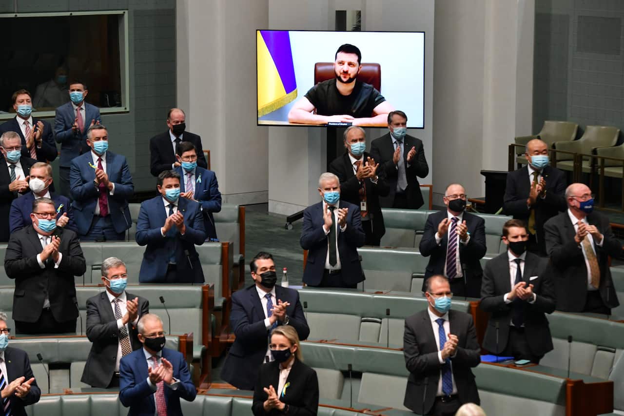 Politicians in the House of Representatives stand and applaud Ukrainian President Volodymyr Zelinskyy, whose image is on a video screen.