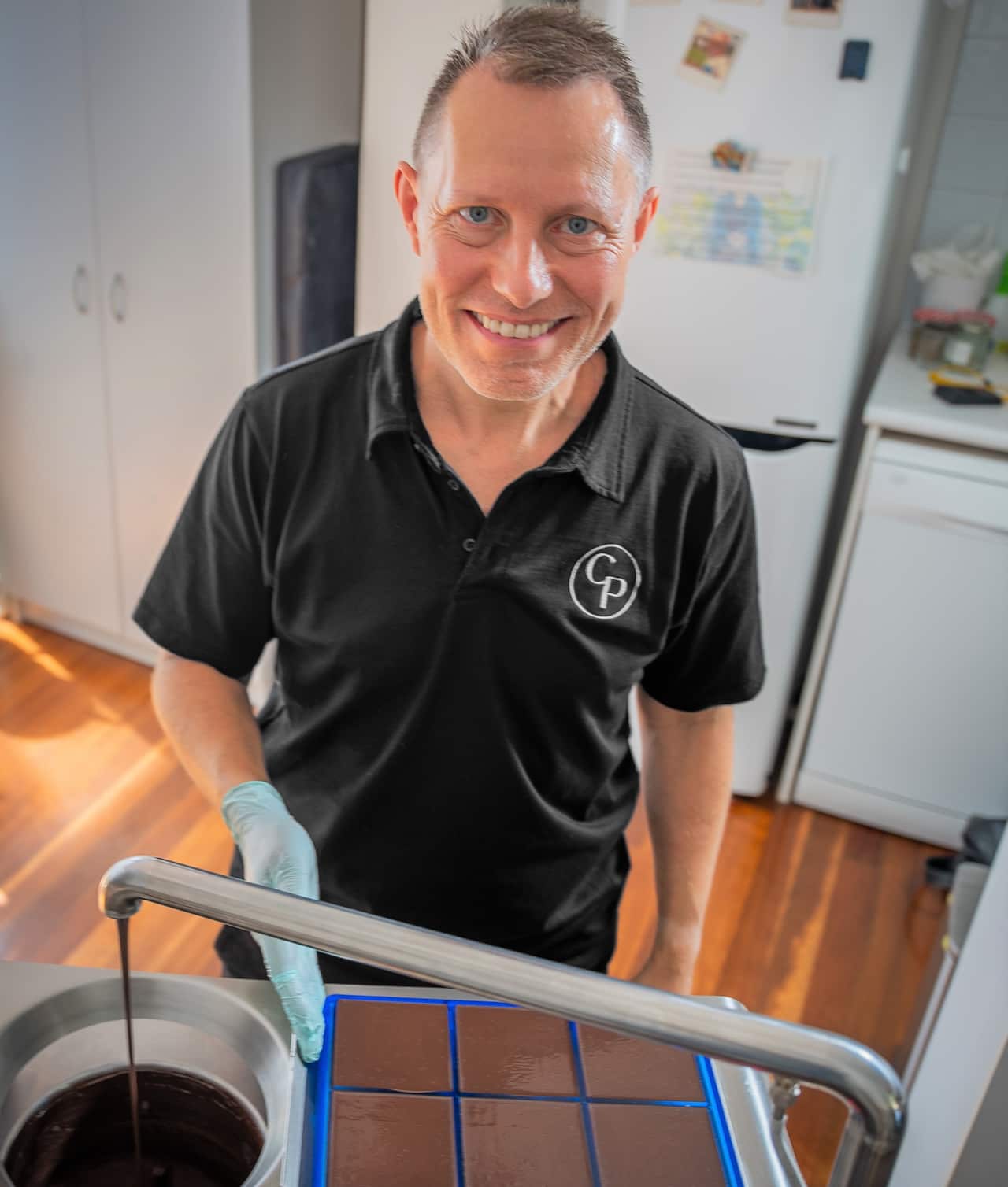 A man in a black T-shirt stands in front of a bench covered in chocolate-making equipment.