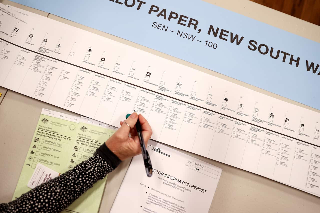 A hand with holding a pen next to a long white piece of paper with printed writing and boxes on it. There is a smaller green piece of paper with writing on it below