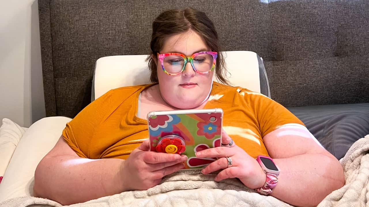 a woman in a yellow t shirt and rainbow glasses is tucked up in bed using a tablet
