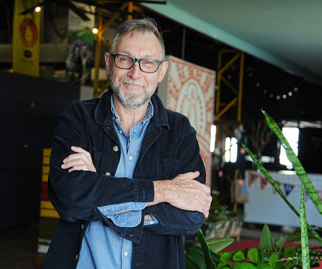 A man in a blue shirt and blazer stands with arms folded against a doorway.