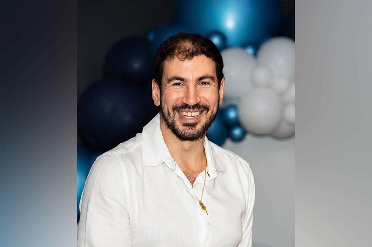 A young white man in a white shirt with short black hair and a beard, smiling. Blue and white balloons are behind him.