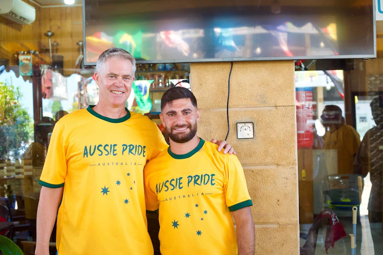 Two men stand arm-in-arm in front of a cafe wearing green and hold Aussie Pride T-shirts.