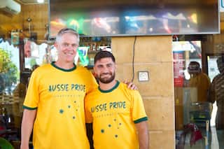 Two men stand arm-in-arm in front of a cafe wearing green and hold Aussie Pride T-shirts.