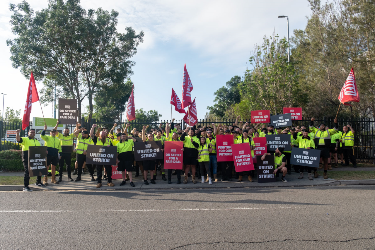 A group of workers in high-visibility clothing holding signs that say "United on Strike," "On Strike for a Fair Deal," and "Fighting for Our Future!"