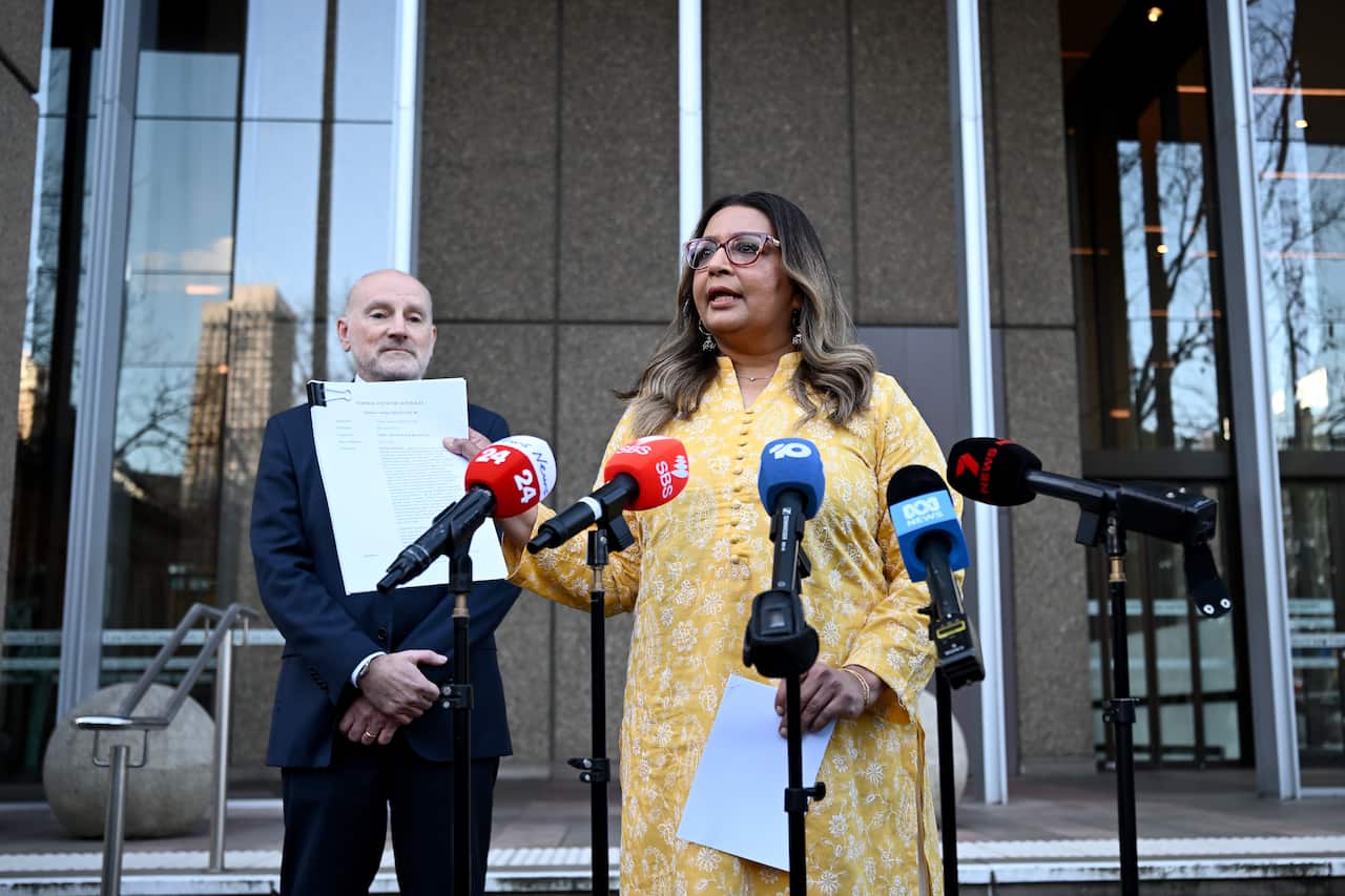 Mehreen Faruqi stands outside of court in a yellow dress holding a piece of paper and speaking into various media microphones.