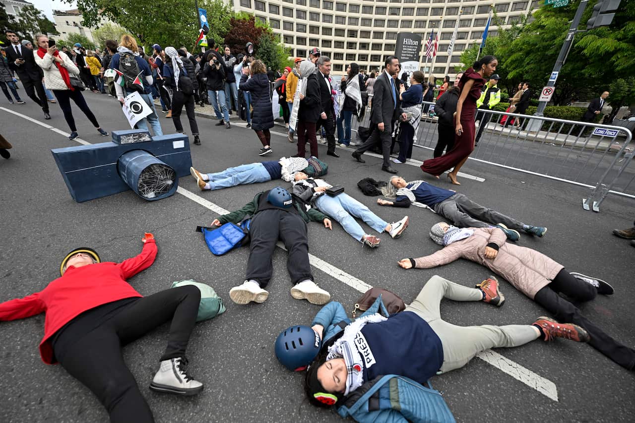 People lying on a road, some of whom are wearing press vests. A large paper mache camera sits behind them