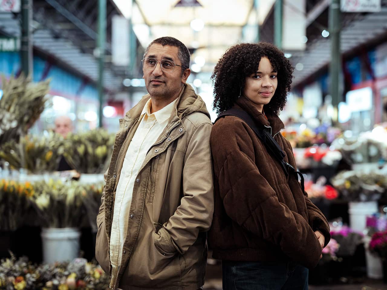 A man and a woman stand back to back in what looks like a flower market. 