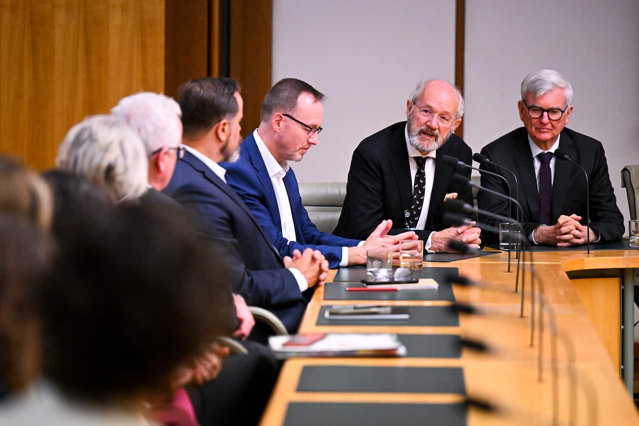 Two men in suits speaking to a number of people sitting in a parliamentary committee room.