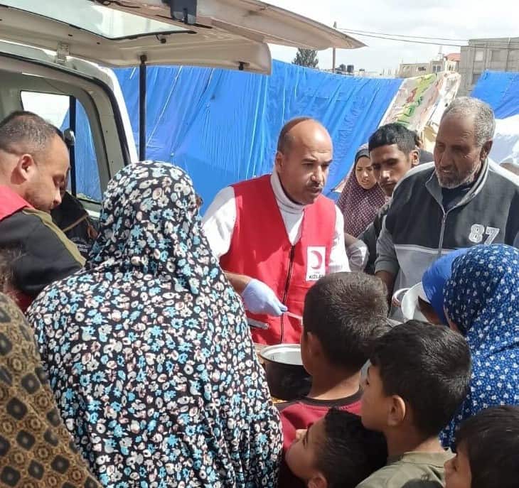 A man in a red vest dishes food out of a pot for the children, women and men in front of him.