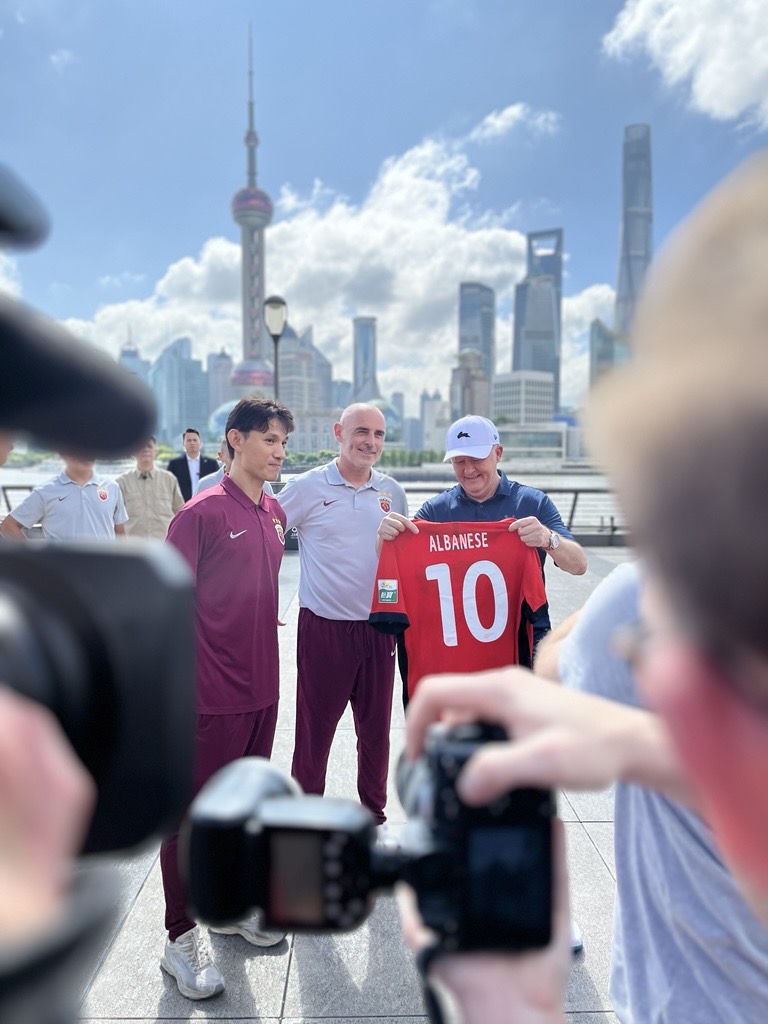 Prime Minister Anthony Albanese wearing a white cap with black rabbit on the front, holding up a vivid red soccer jersey with the number 10 and ALBANESE in white lettering on the back. He is standing two other men.