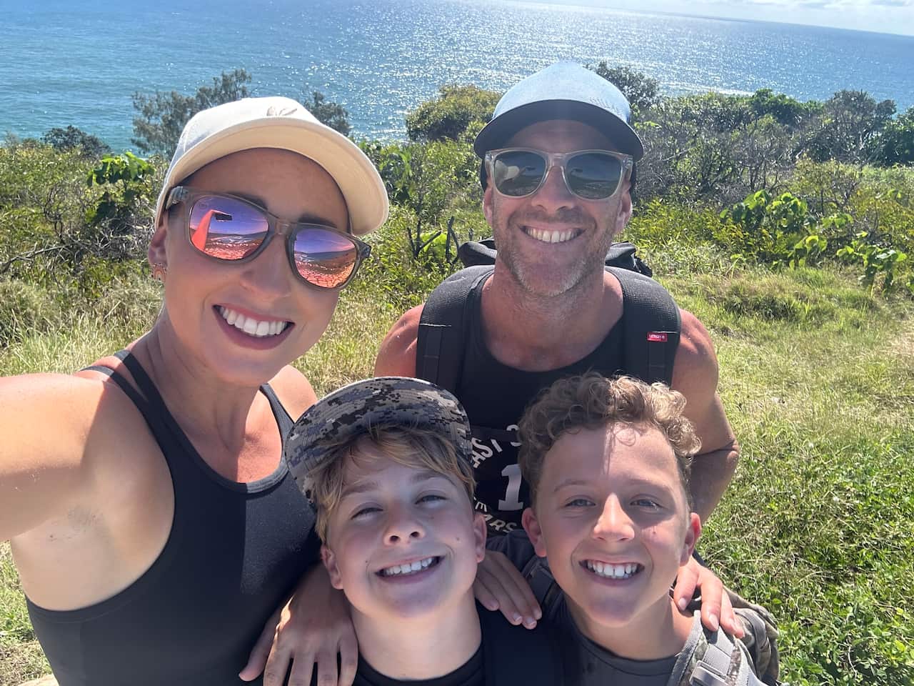 A woman, man and two young boys smile for a selfie taken by the woman in front of a glimmering blue ocean.
