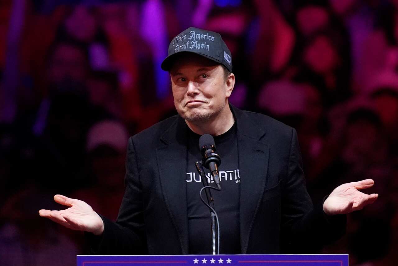 Elon Musk shrugs at a lectern, wears a black 'Make America Great Again' cap, and speaks into a microphone.