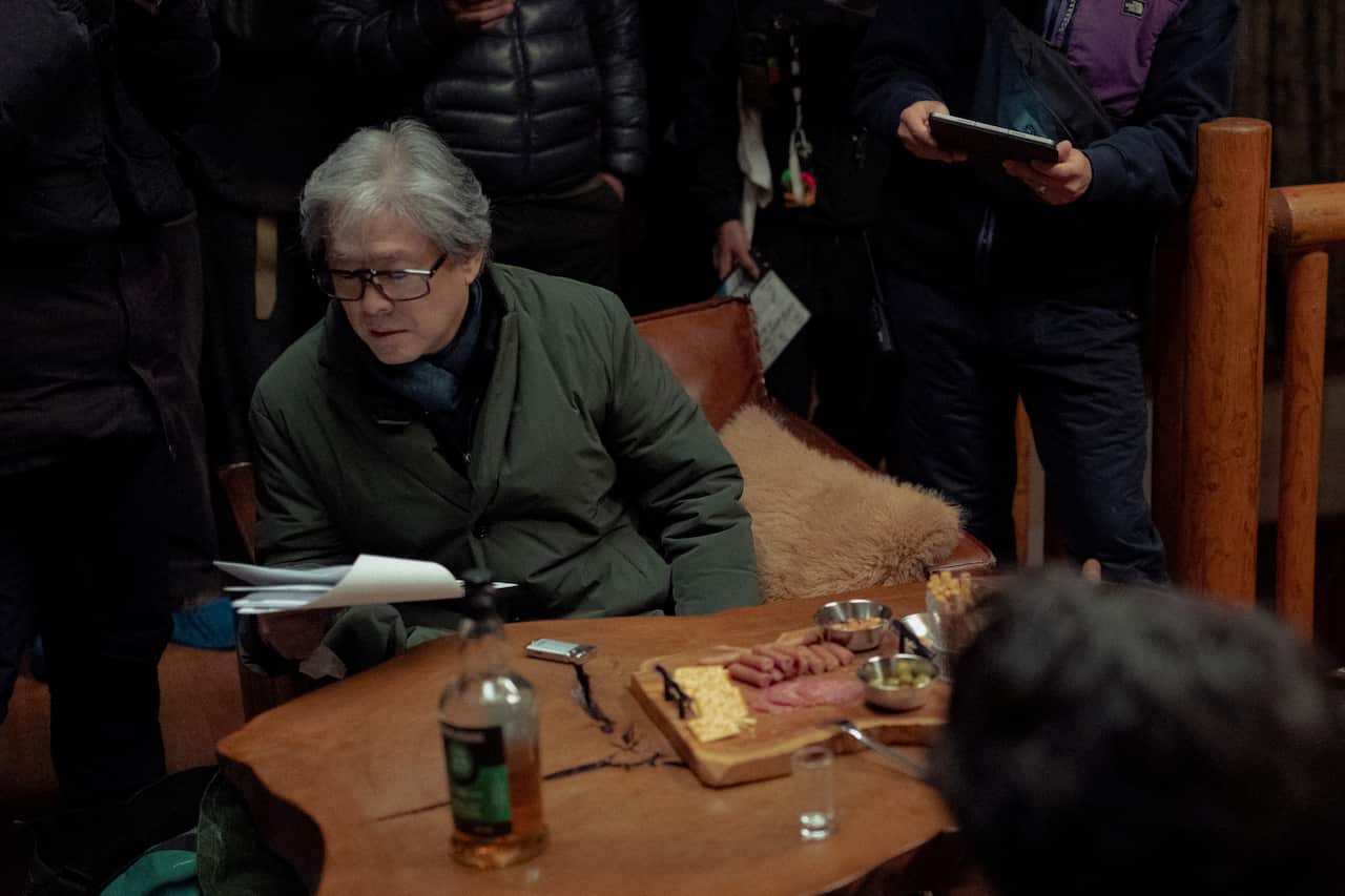 A behind the scenes shot of Park Chan-wook on a film set. He is holding a script and there is a cheese board in front of him.