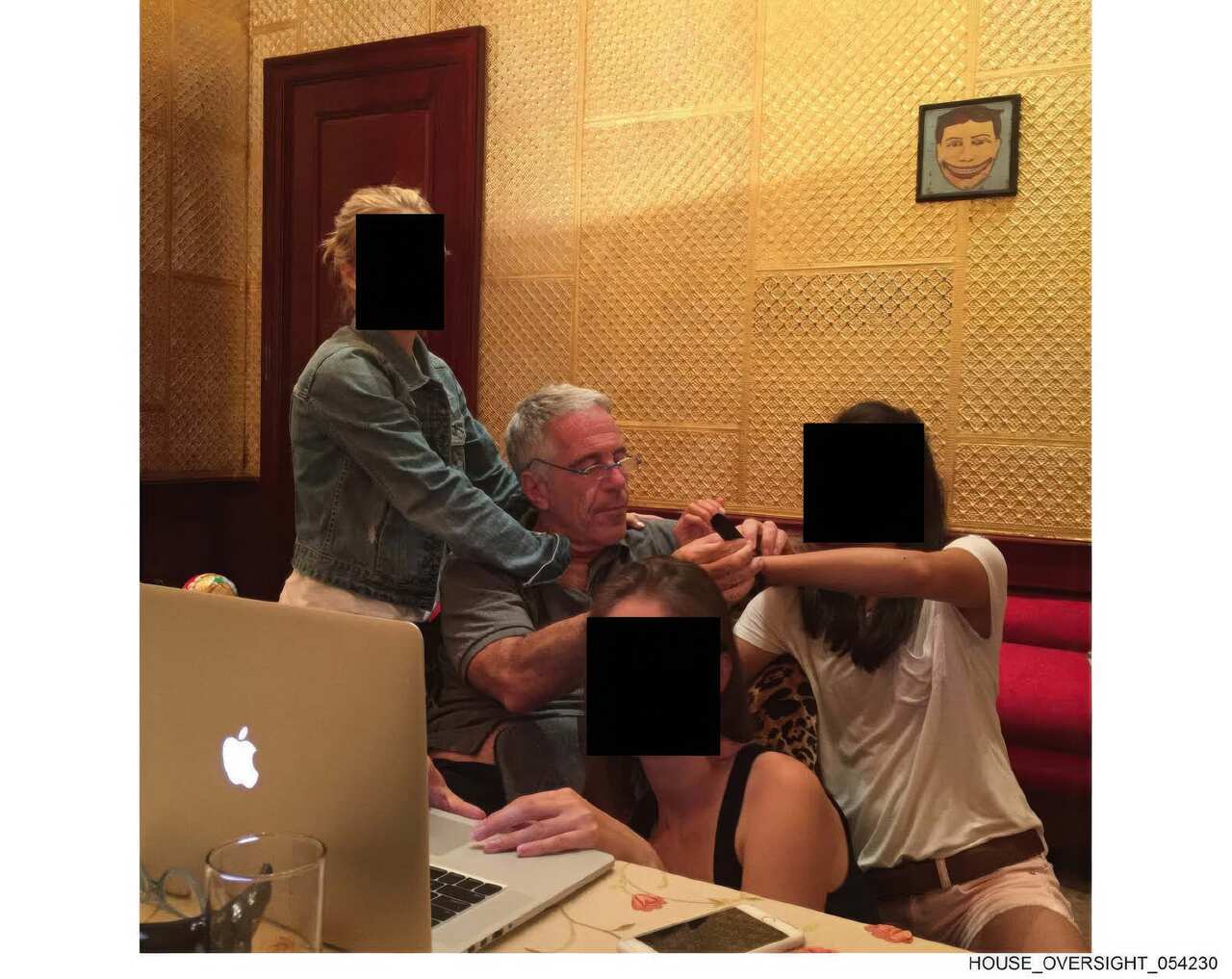 Jeffrey Epstein sits at a table with three young women, whose faces are obscured by black squares, in a room with patterned gold walls and a framed drawing of a smiling face.