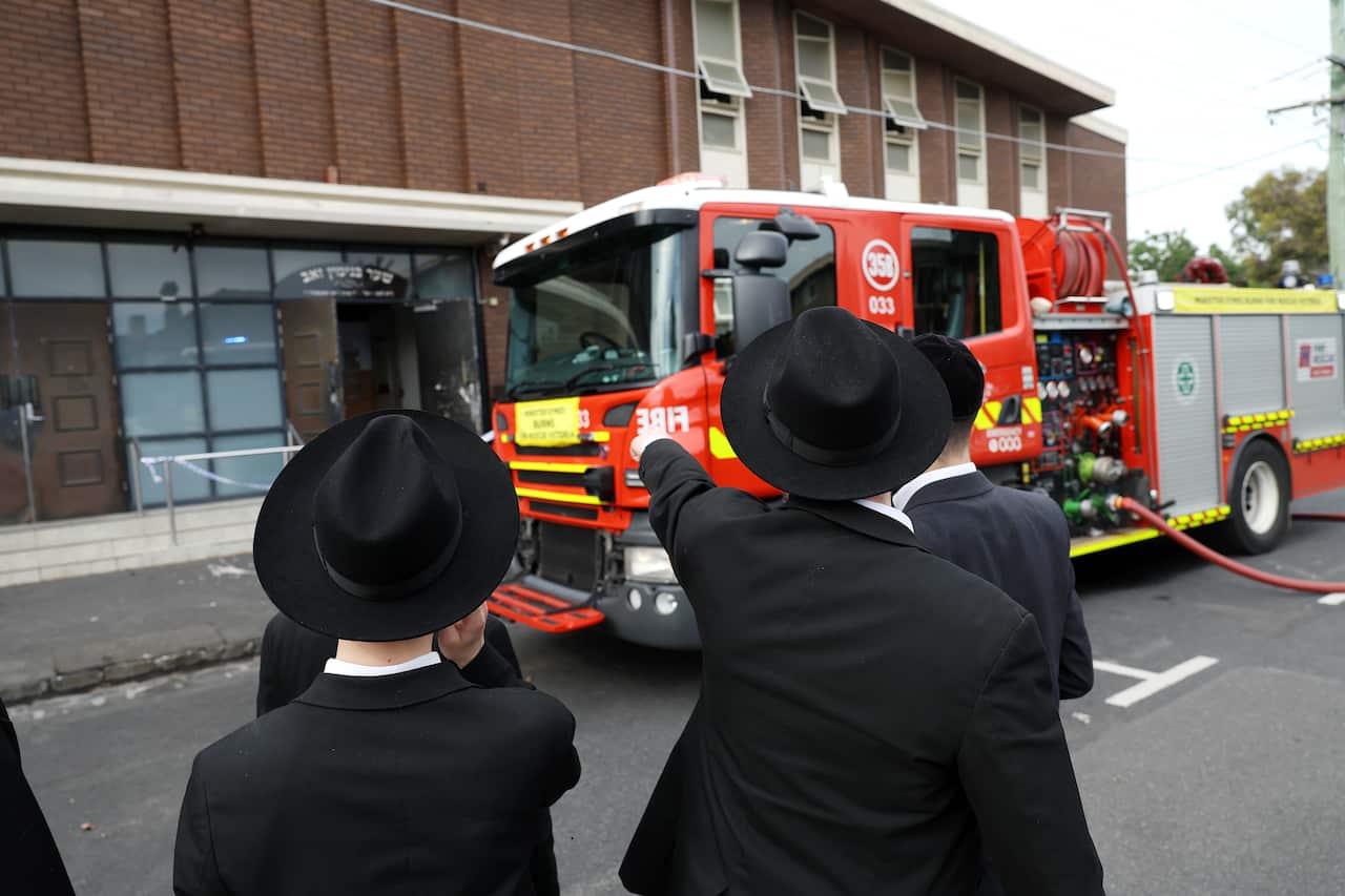A man in a black suit and hat stands beside a woman in similar attire, pointing at a building with a fire truck parked in front.