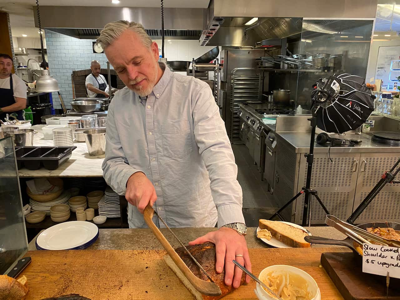 A man wearing a white shirt is using a knife to cut a loaf of bread placed on an island in a professional kitchen.