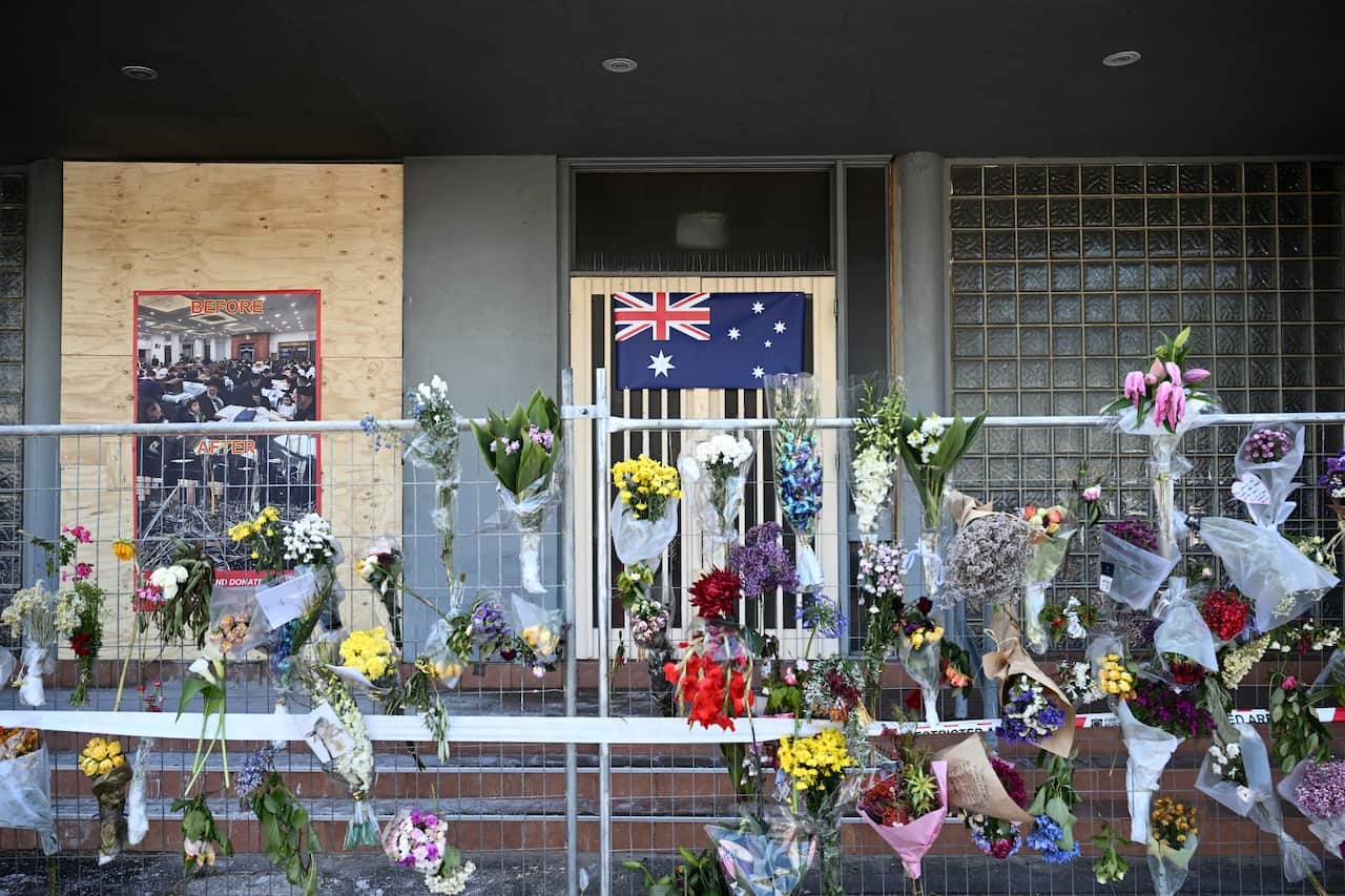 A fence in front of a building with boarded-up walls is covered in bouquets of flowers and an Australian flag.