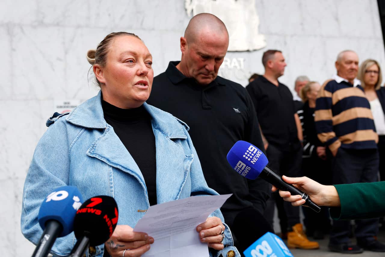 A middle-aged white woman wearing a blue coat speaks to the media outside a courtroom. Her husband stands beside her. 