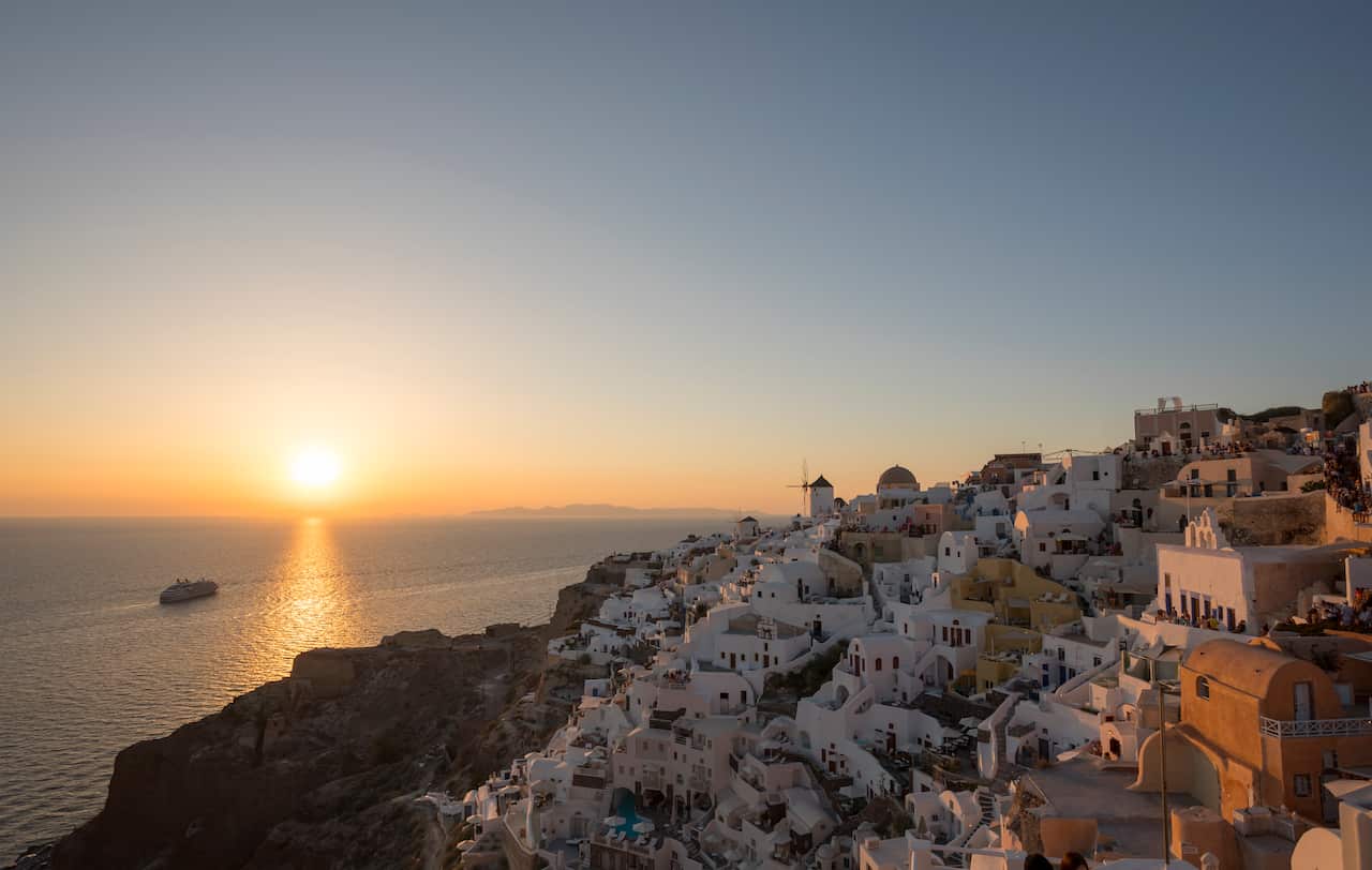 The sun sets over the sea. A village of white houses set against the cliffs overlooks the ocean. 