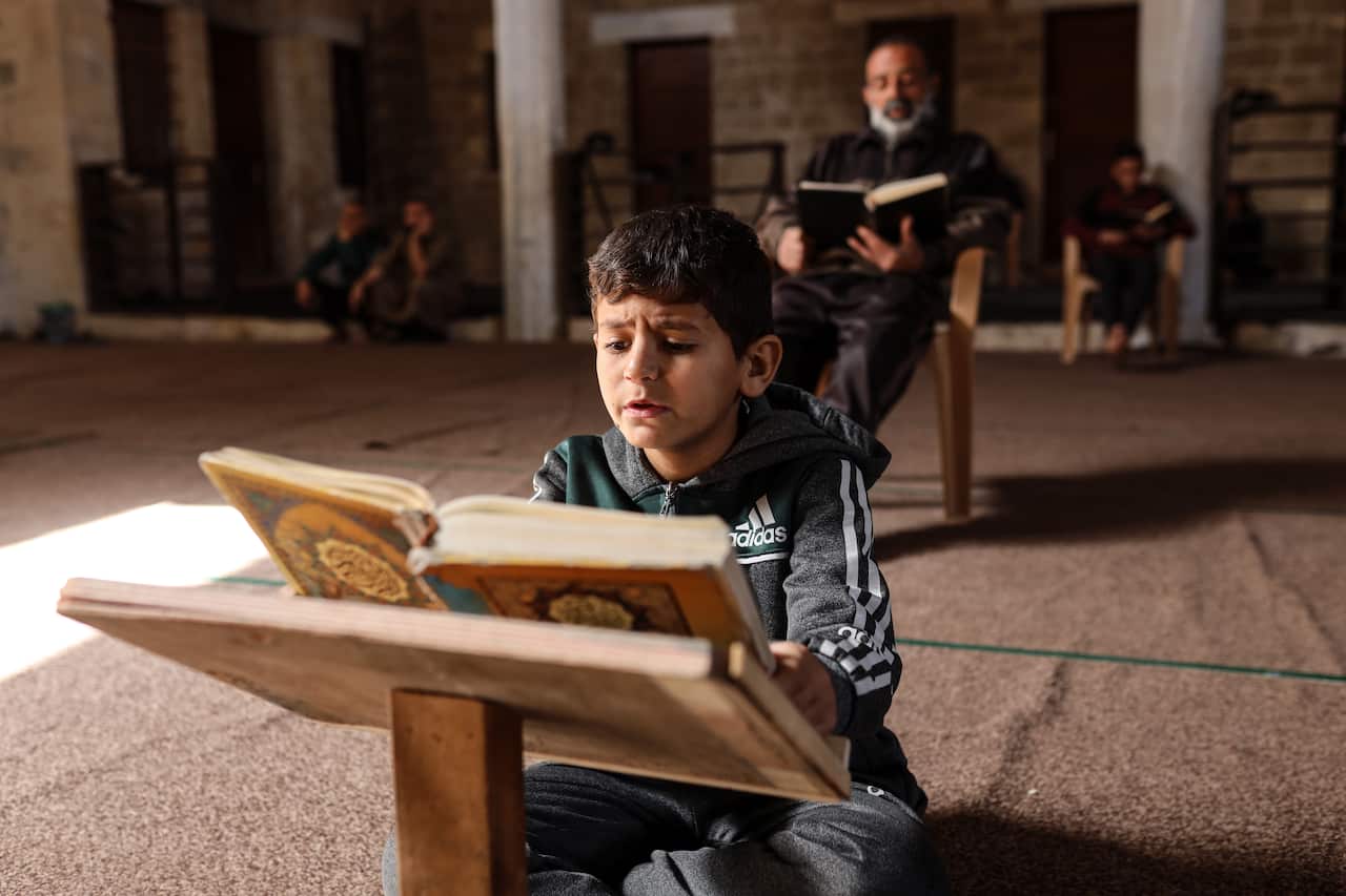 A little boy sitting on the floor reading a Quran, men reading behind him.