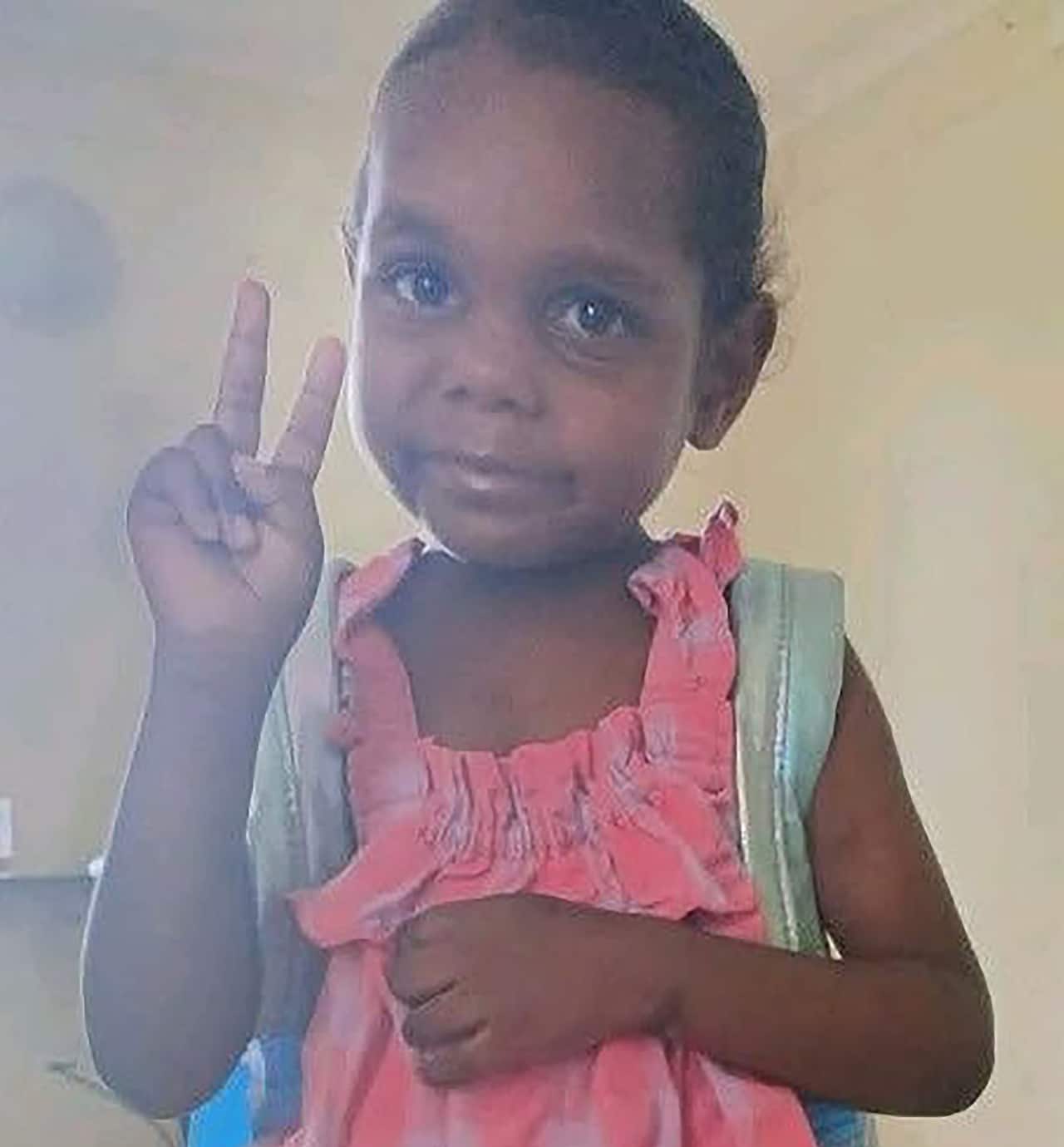 A young child with dark hair pulled back wears a pink ruffled dress and a light blue backpack while making a peace sign with one hand.