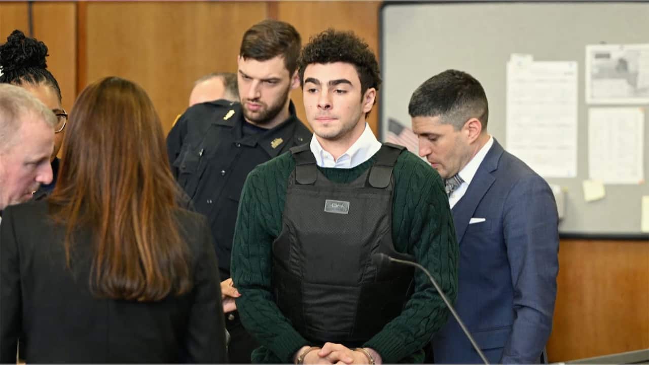 Luigi Mangione in court. He stares ahead, wearing a green knit jumper and a bullet-proof vest. He is surrounded by New York City police and lawyers.