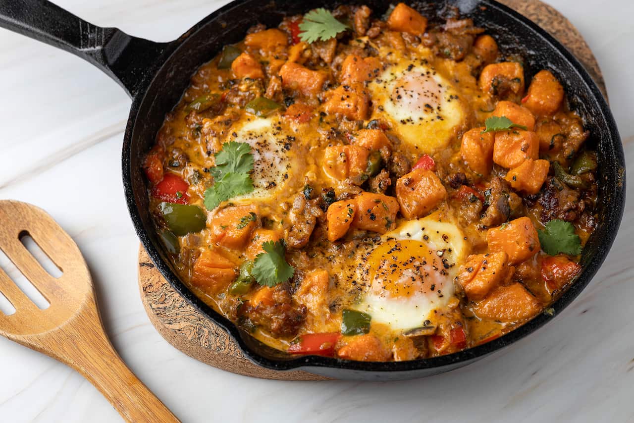 A cast iron skillet sits on a matt on a white surface with a slotted wooden spoon beside it. The pan has a stewish mixture of sweet potato, sausage, scattered herbs and two eggs that have been cooked in the mix.  