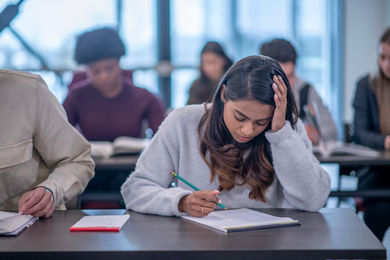 A female university student has one hand on her head while using the other to write an exam, with other students visible in the background.