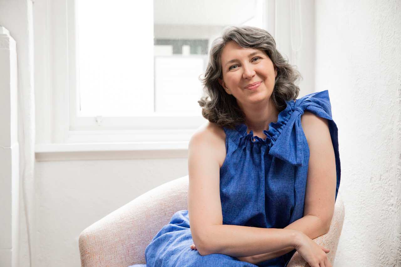 A woman in a blue dress and silver hair smiles while seated in an armchair 