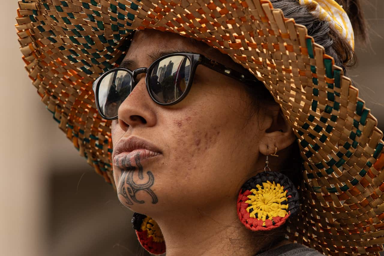 A person wearing a large straw hat, sunglasses and an earring that depicts the Aboriginal flag.