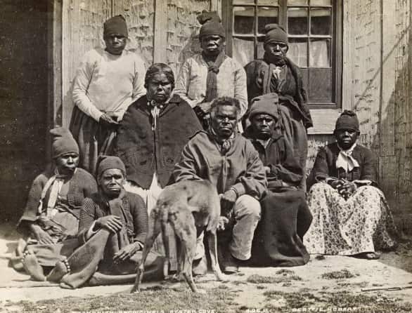 A black and white image of nine Aboriginal people dressed in western clothes sitting outside a simple house