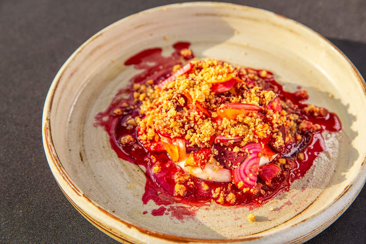 A round pottery bowl in cream tones holds a serve of a vibrant pink beetroot 'pudding', topped with a golden crumble and sitting in small puddle of pink juices.  