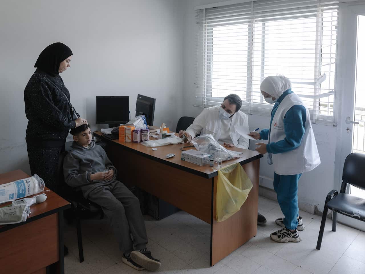 A doctor sits at a wooden table in a clinic with a young patient across from him. A woman in a burqa is standing beside the patient, while a nurse stands next to the doctor.