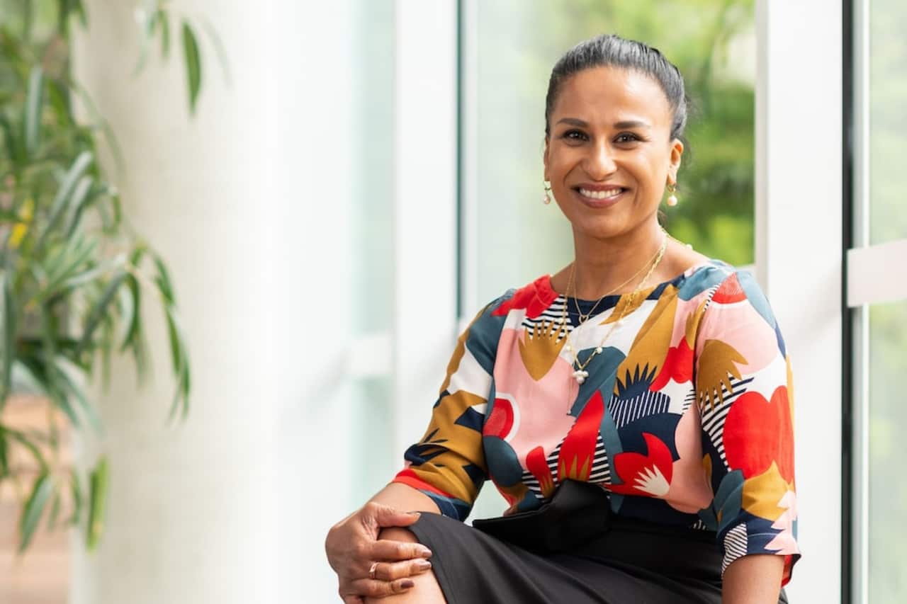 Madhavi Nawana Parker wearing a colourful floral print top and black skirt. She is seated as she smiles at the camera.