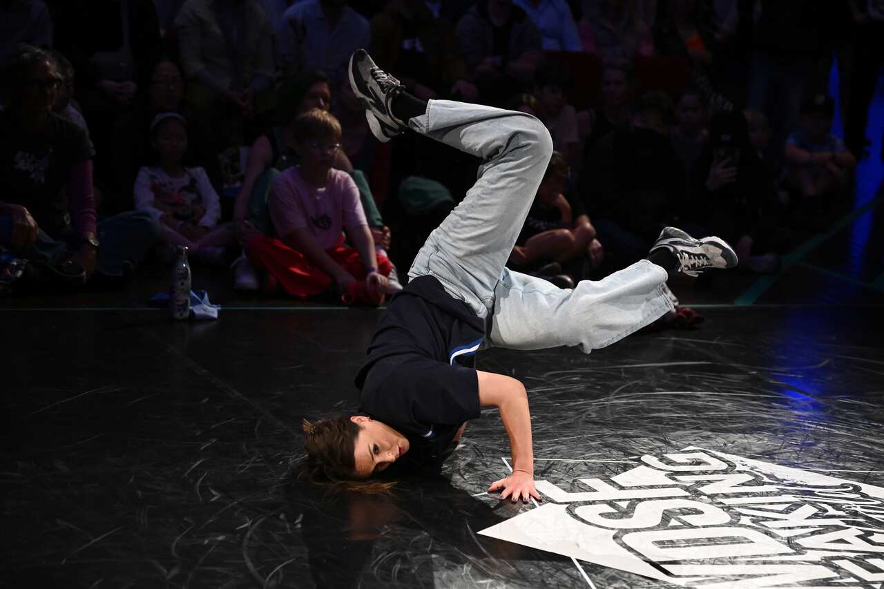 A woman in grey tracksuit pants breakdances on a black floor with spectators in the background. 