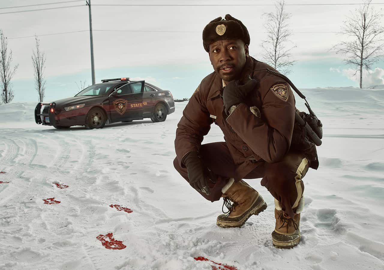A man in a brown uniform crouches in the snow beside a trail of bloody footprints. 
