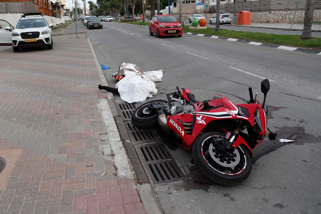A corpse covered with a white sheet lies beside a motorbike on its side.