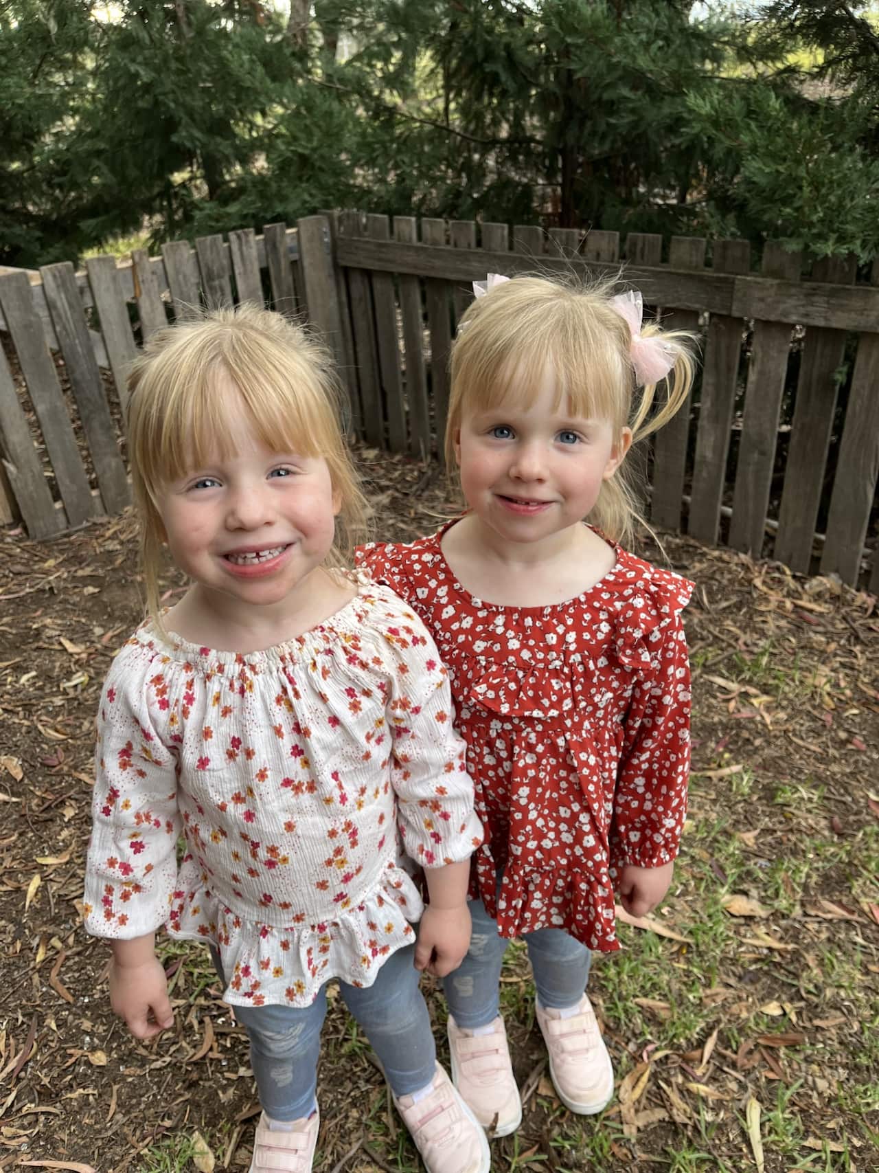 Two young girls wearing floral tops and blue jeans smiling in the corner of a backyard