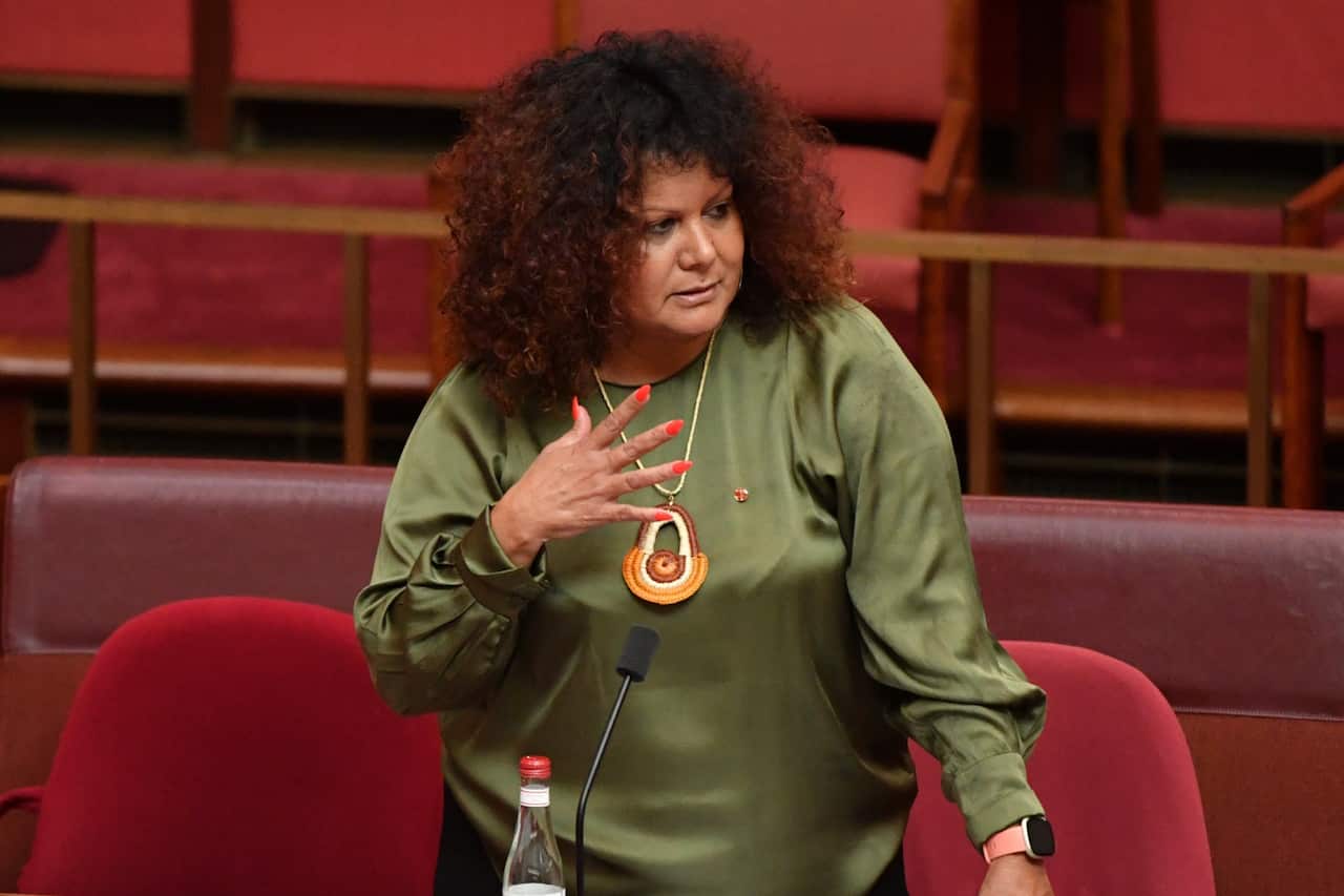 A woman stands surrounded by the red seats of the Senate chamber. She wears a green shirt with an ornate necklace.