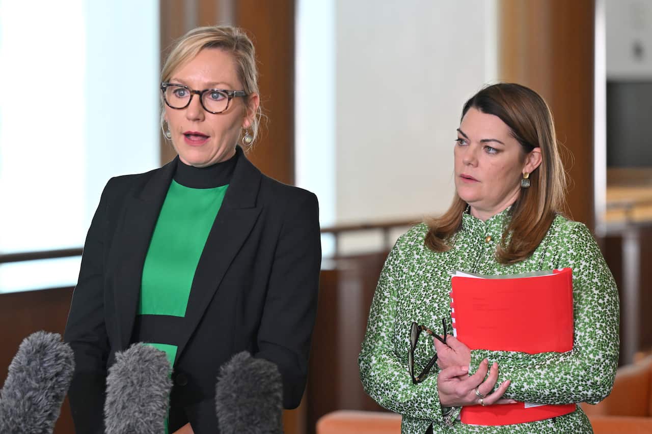 Woman in green speaks into microphone, accompanied by woman holding a red binder.