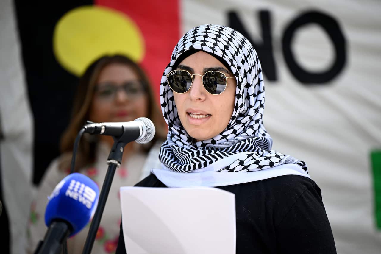 A woman wearing black sunglasses and a white and black patterned headscarf talks into a microphone.