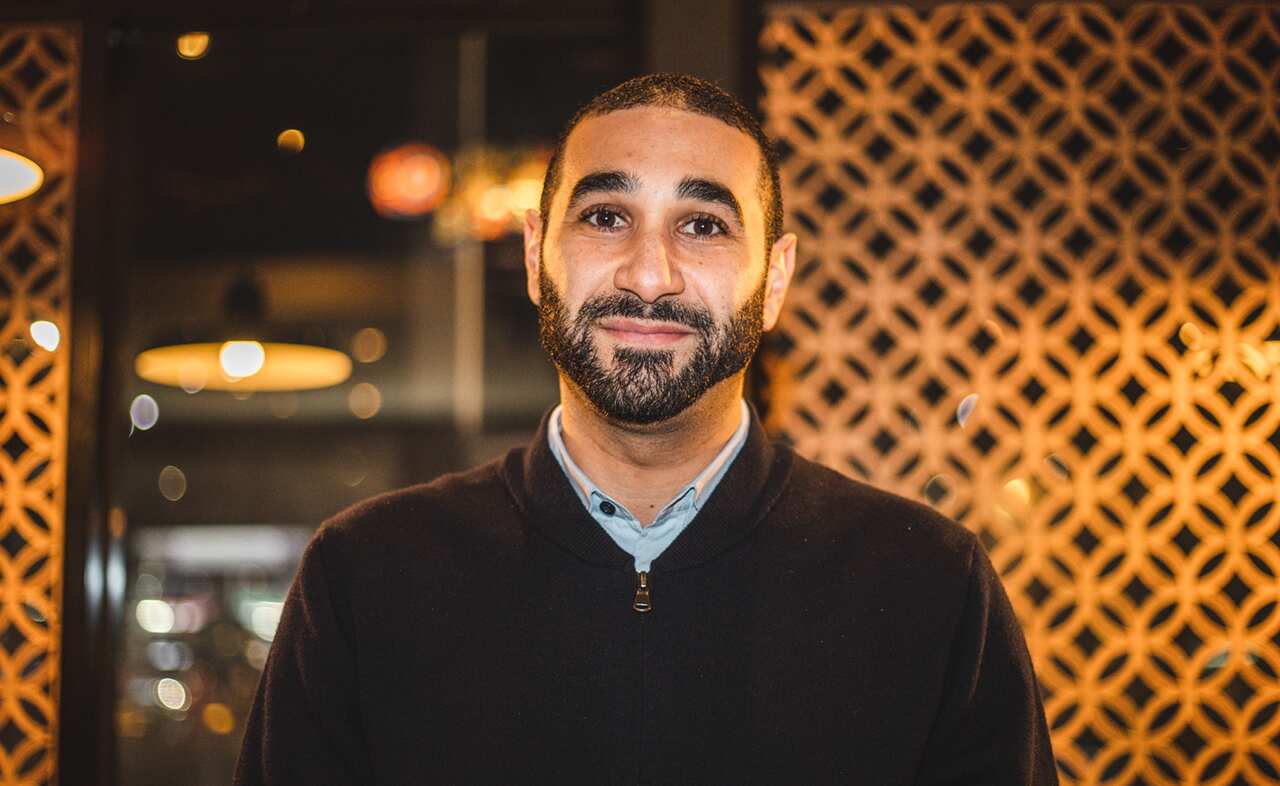 A gently smiling man with short hair and a short beard stands in front of a patterned orange panel.