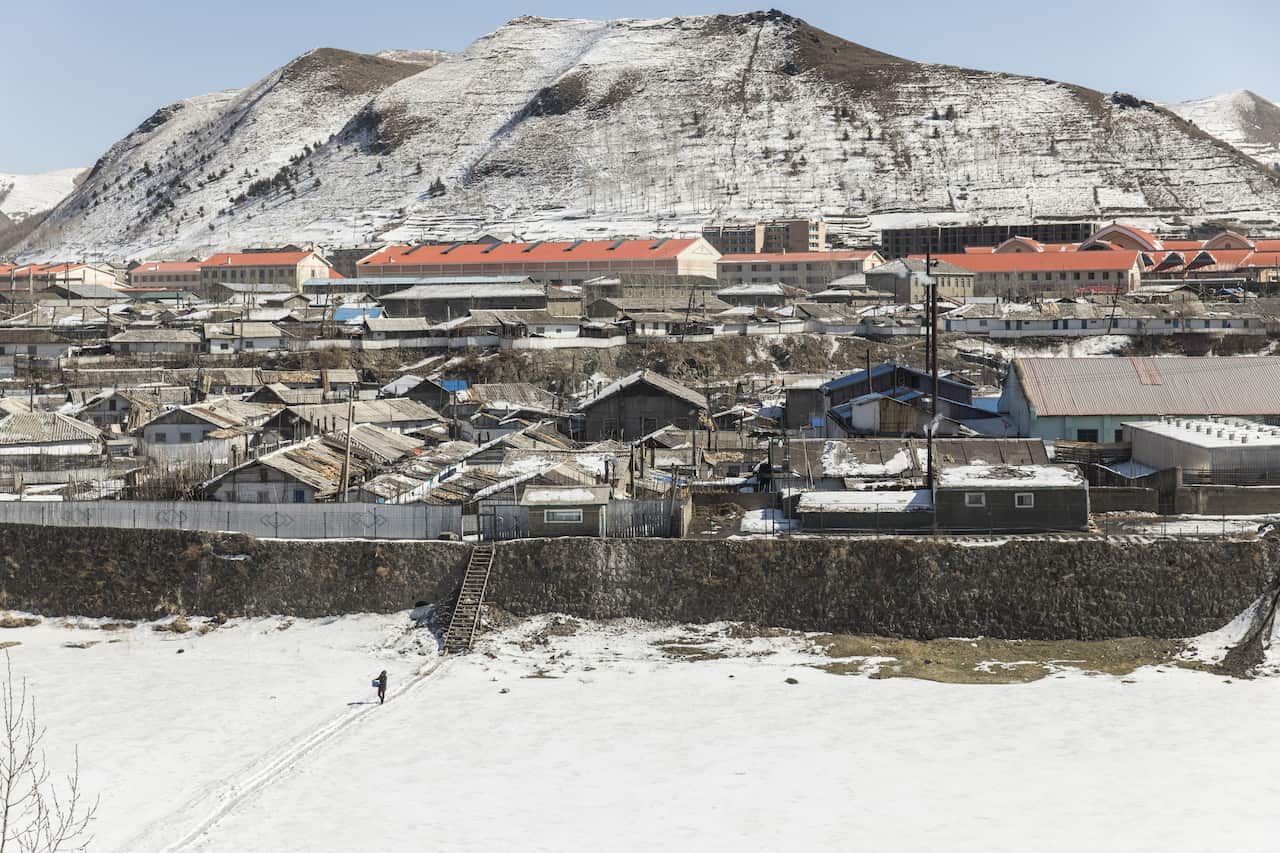 A city with low buildings covered in snow sits below a snow-topped mountain. In the foregroun, a figure carrying a bucket walks along a snowy plain. 