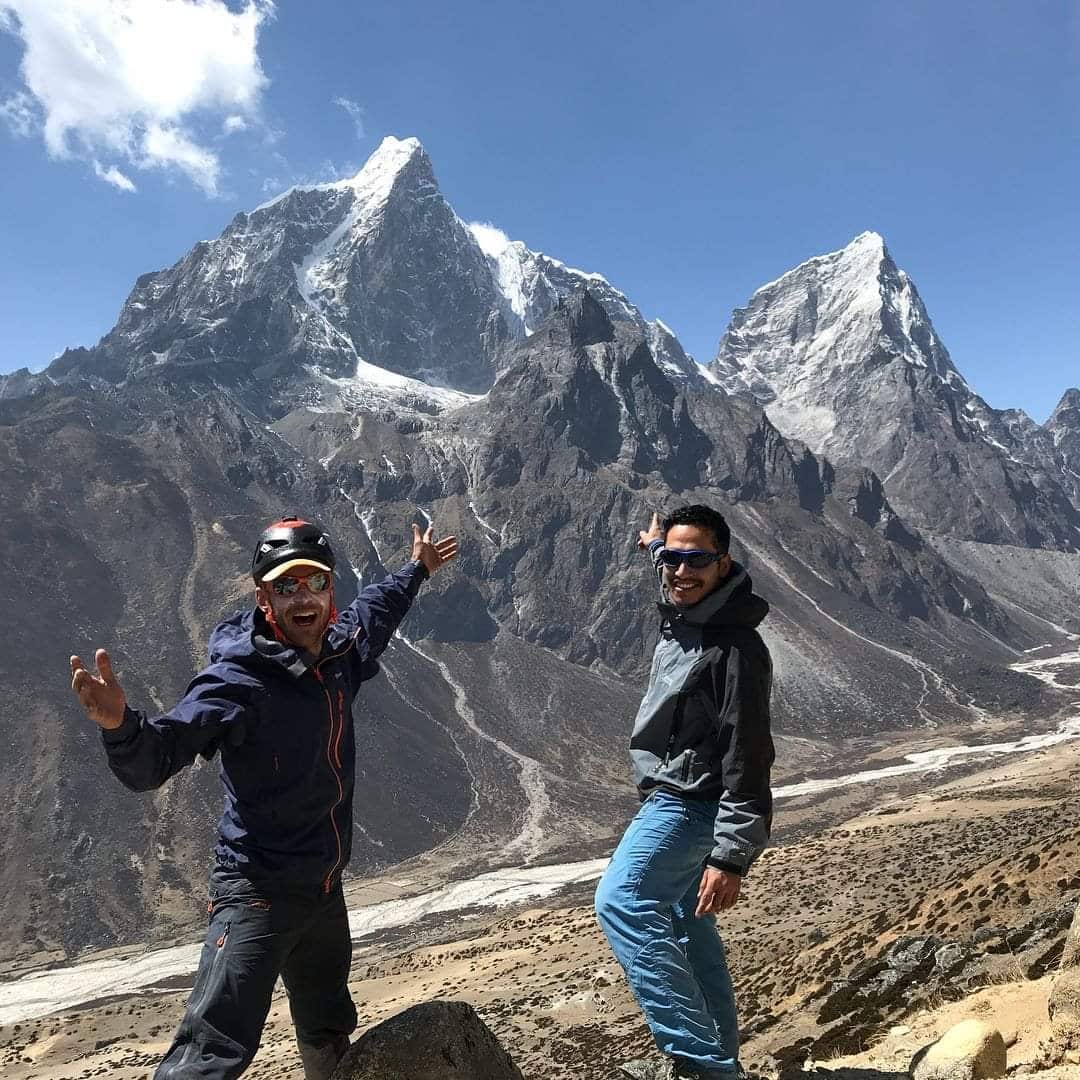 Two men smile and point to a mountain. 