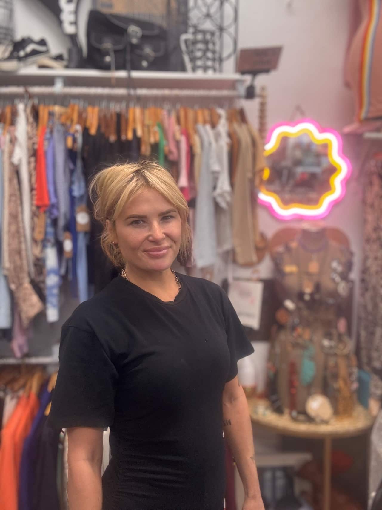A woman with blonde hair is wearing a blue t shirt smiling with clothing racks behind her