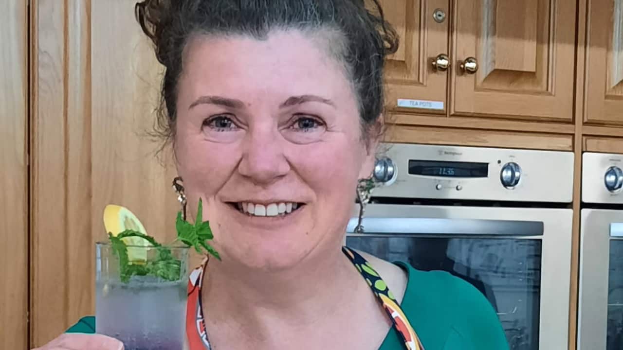 a middle age woman in an apron smiles in a kitchen holding a mocktail up to the camera
