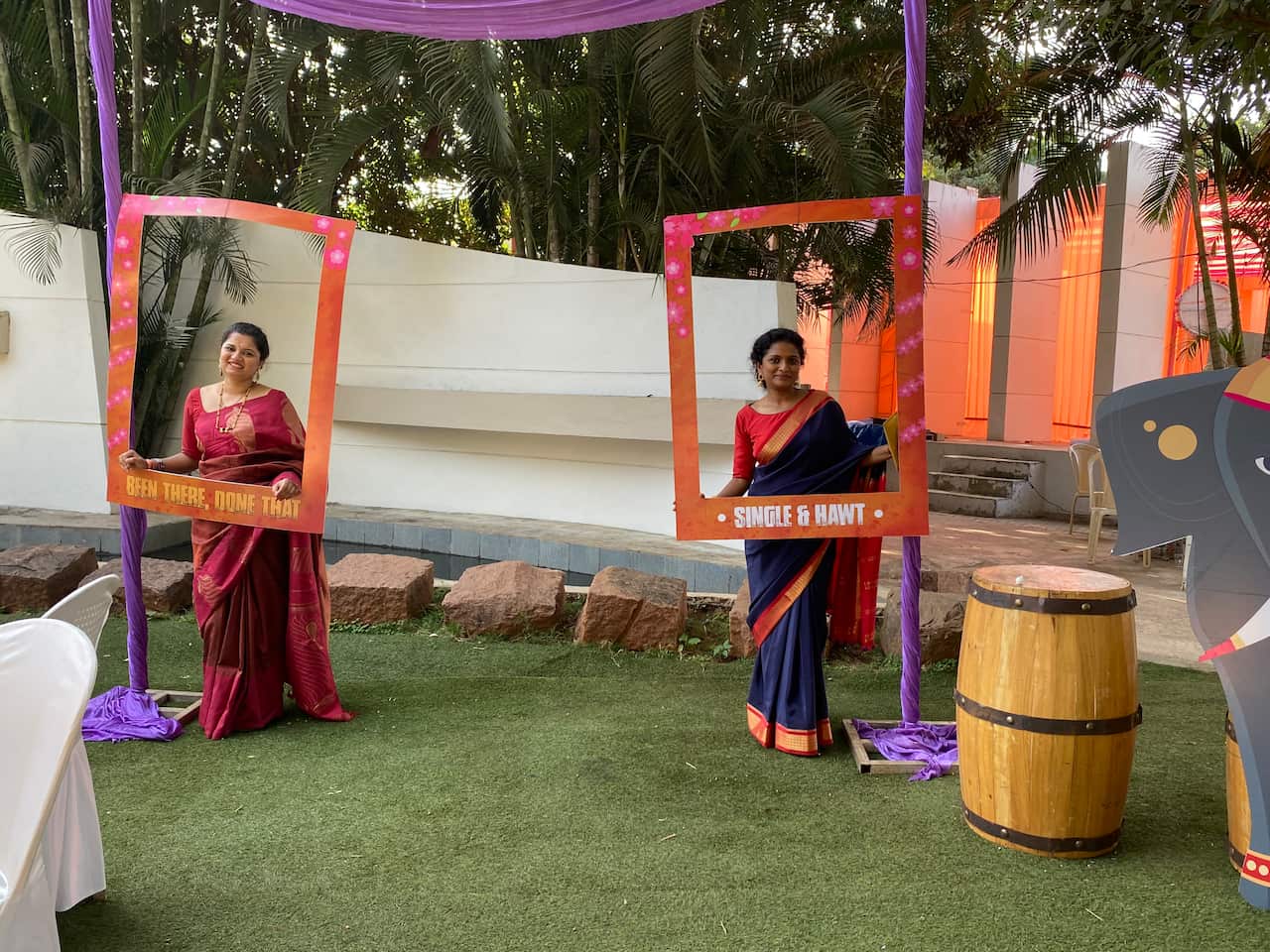 Two Indian women in saris stand in a grassed courtyard at a wedding, one holding a frame that says 'Been there, done that', another holding a frame with the words, 'Single & hawt'.