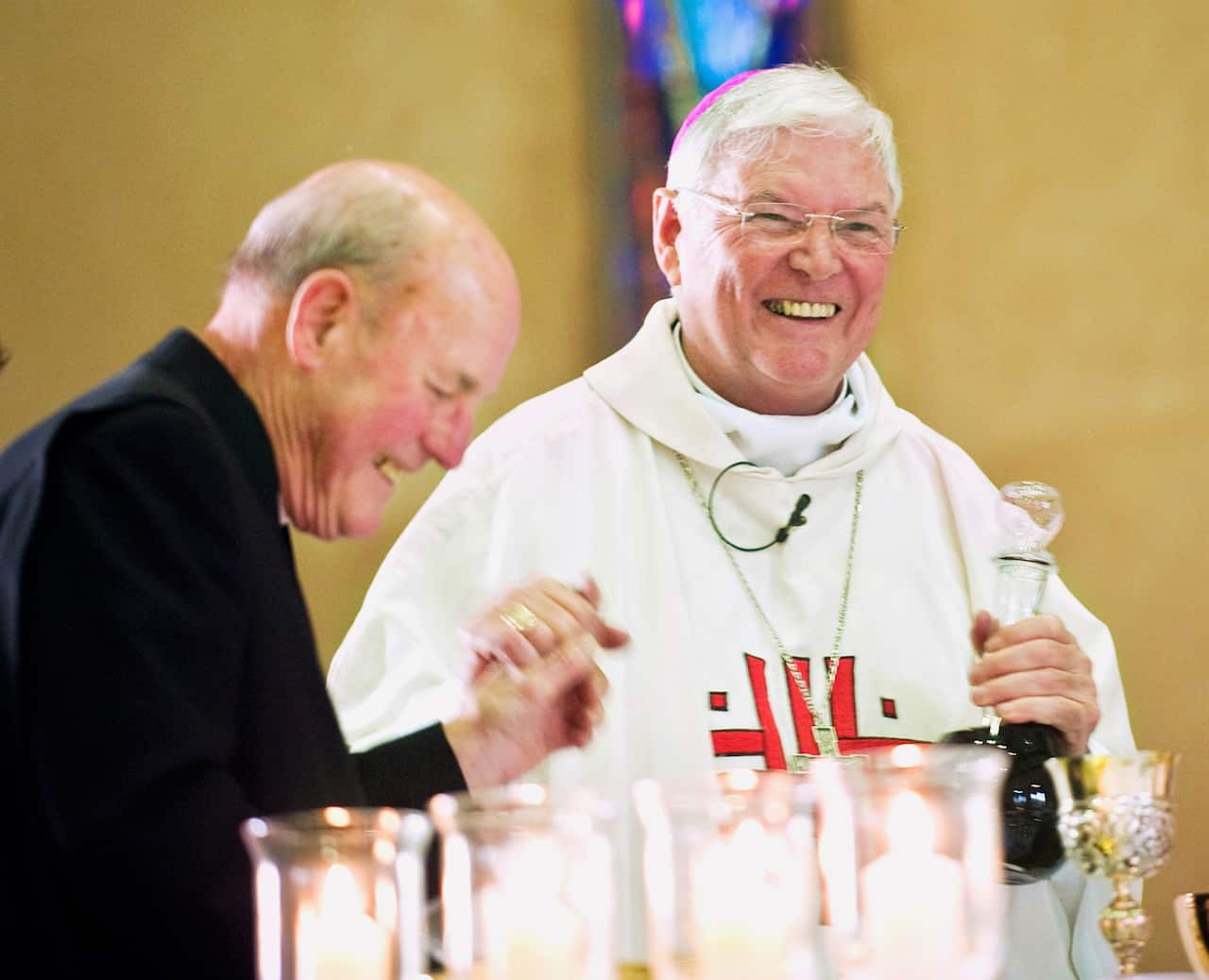 A Jesuit bishop in formal robes stands in a church with another priest nearby. They are both laughing.