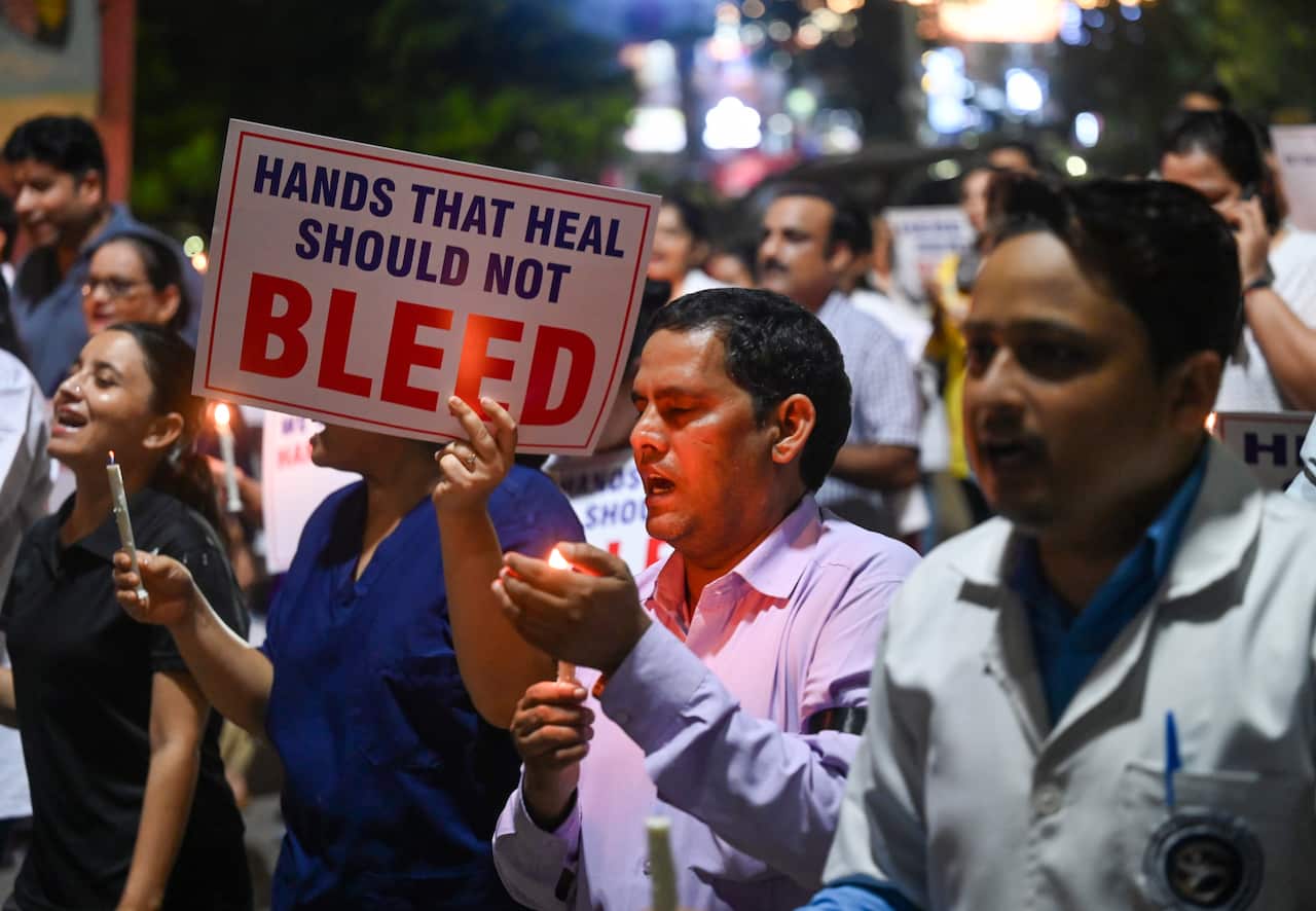 A group of people protesting, some with small candles. One person is holding up a sign that reads: "Hands that heal should not bleed."